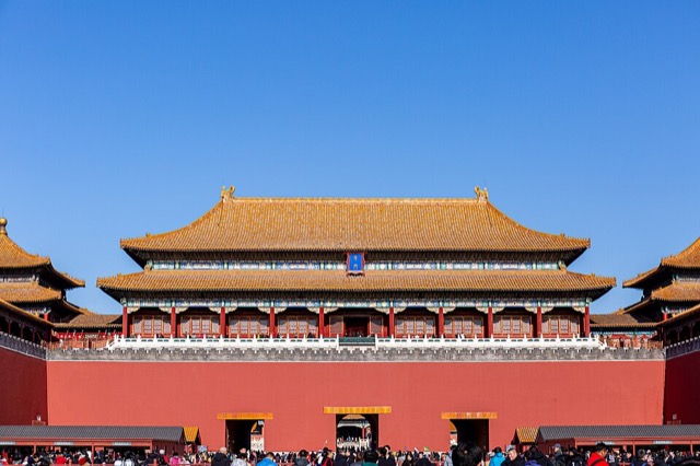 La porte du Midi vue de face sous un ciel bleu : le pavillon central à double toit doré domine un massif de brique rouge percé de trois arches, avec les deux ailes en retour de chaque côté.