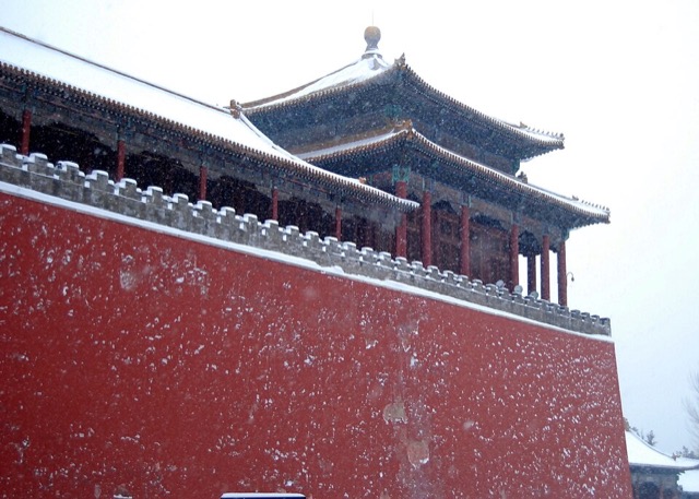 Sous la neige qui tombe, un pavillon d’angle de la porte du Midi, au toit pyramidal coiffé d’une boule dorée, surmonte le mur rouge crénelé.