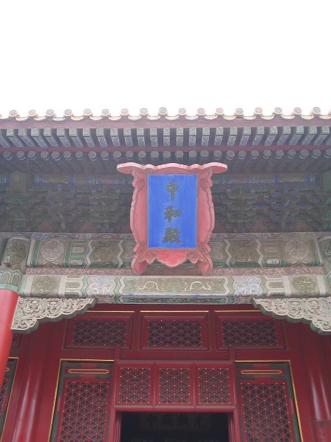Gros plan sur le cartouche bleu au nom de la salle de l’Harmonie parfaite, suspendu sous l’avant-toit au-dessus de portes rouges à claire-voie.