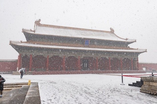 La salle de l’Harmonie préservée sous une chute de neige, ses deux toits blanchis au-dessus des colonnes rouges, avec un visiteur sous un parapluie sur le parvis enneigé.