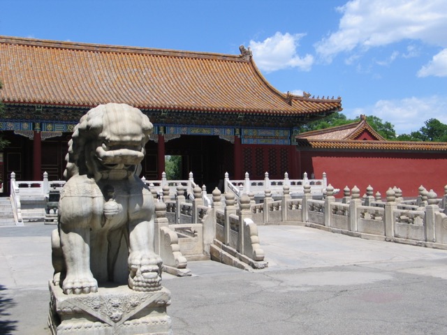 Un lion de pierre sur son socle sculpté, la patte posée sur une boule, devant les balustrades de marbre et la porte de la Prouesse militaire, sous un ciel bleu à nuages.