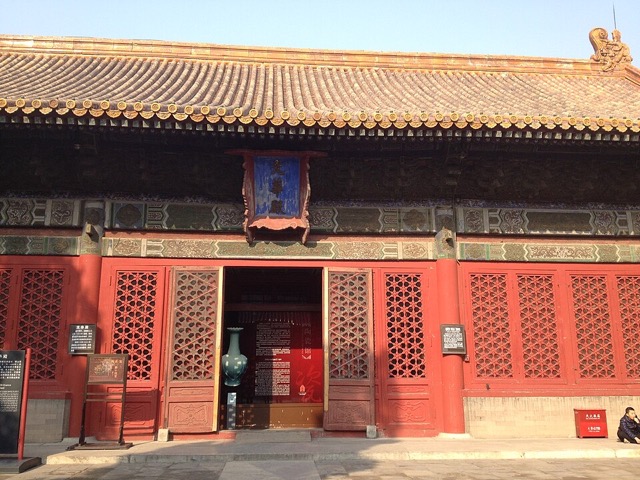 Gros plan sur la façade de la salle de la Gloire littéraire : le cartouche bleu au nom de la salle sous l’avant-toit, et des portes rouges à claire-voie dont l’une, ouverte, laisse voir un grand vase.