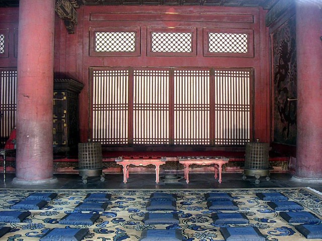 Intérieur aux colonnes rouges et cloisons à claire-voie, deux petits bancs laqués rouge et des rangées de coussins bleus sur un tapis à fleurs.