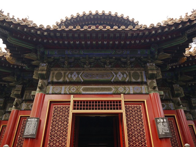 Détail de la façade du pavillon des Dix mille printemps en contre-plongée : consoles peintes de bleu et de vert, portes rouges à claustra et panneaux explicatifs.