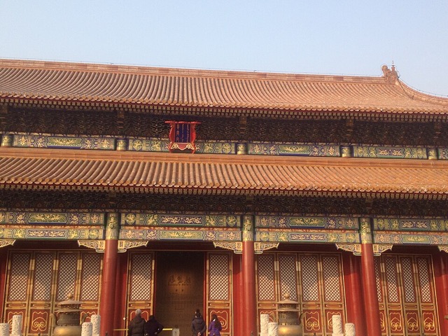 Façade d’une salle à double avant-toit de tuiles jaunes, vue de face en contre-plongée, avec ses colonnes rouges et ses portes à claire-voie.