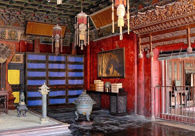 Une pièce rouge et or aux lanternes suspendues, avec des cloisons bleues à panneaux, un rideau jaune près d’un siège et un brûle-parfum émaillé.
