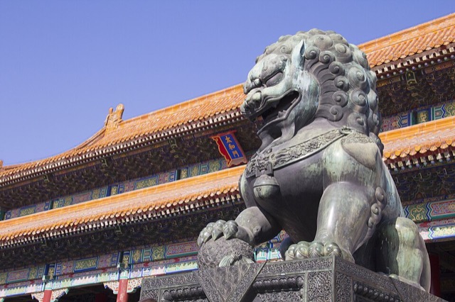 Gros plan sur le lion de bronze qui garde la porte de l’Harmonie suprême, la patte posée sur une boule, devant la façade et le cartouche bleu au nom de la porte.
