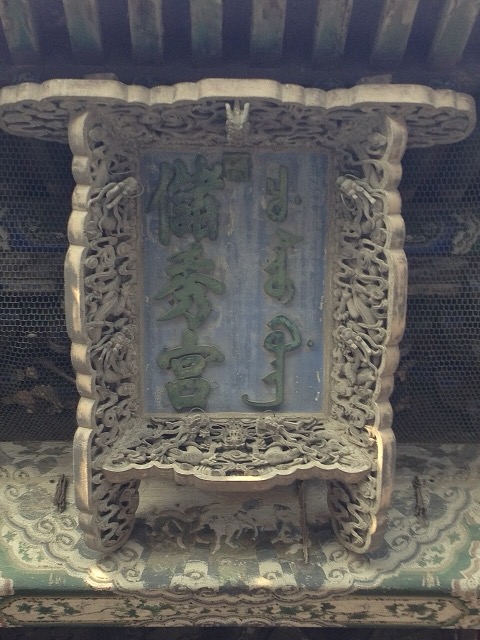 La plaque du palais de l’Élégance rassemblée : caractères verts sur fond bleu, en chinois et en mandchou, dans un cadre de bois ajouré sculpté de dragons et de nuages.