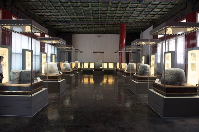 Intérieur d’une salle au plafond à caissons peints, où des blocs de pierre arrondis sont exposés dans des vitrines alignées.