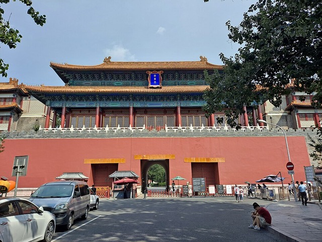 Porte de la Prospérité de l’ouest vue de face : mur rouge à trois arches sous un pavillon à double avant-toit, voitures garées devant.