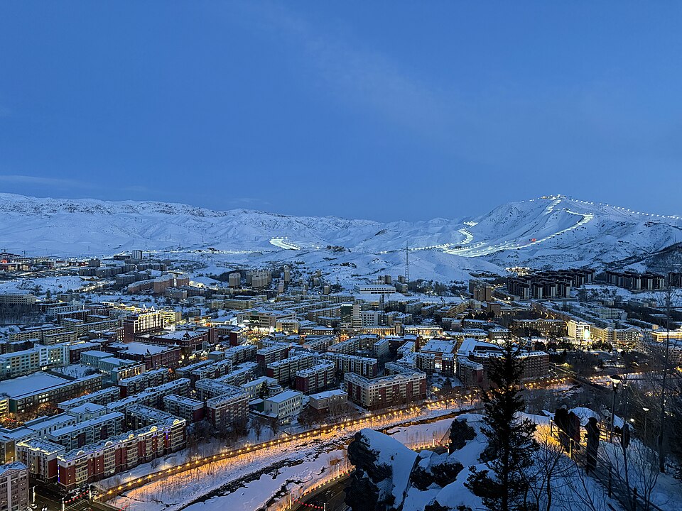 Une ville enneigée s'allume au crépuscule dans une cuvette de montagnes blanches ; sur le versant du fond, les pistes éclairées d'un domaine skiable dessinent de longs traits lumineux.