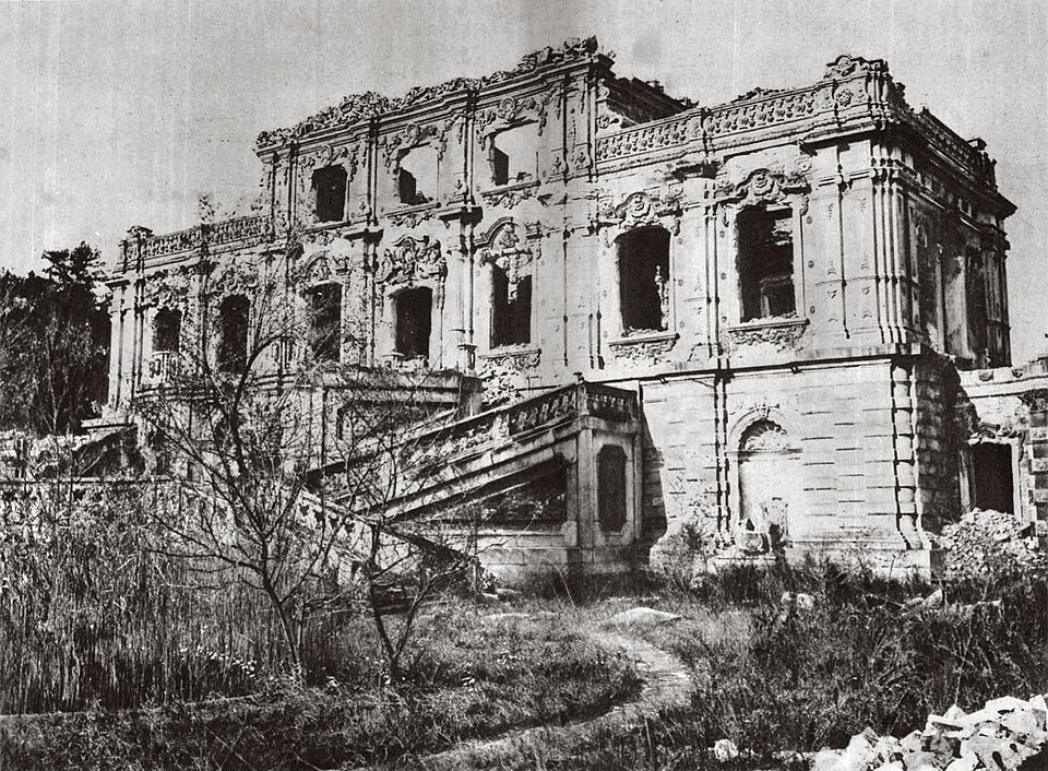 Photographie ancienne des ruines d'un palais de pierre à l'européenne, colonnes brisées et façade éventrée, envahies d'herbes