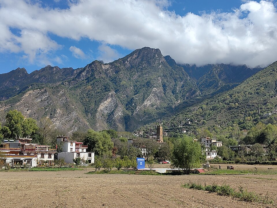 Des maisons blanches à toit plat s'étagent sur un replat au pied d'une haute montagne grise ; devant elles un champ nu, et de gros nuages accrochés à la crête.
