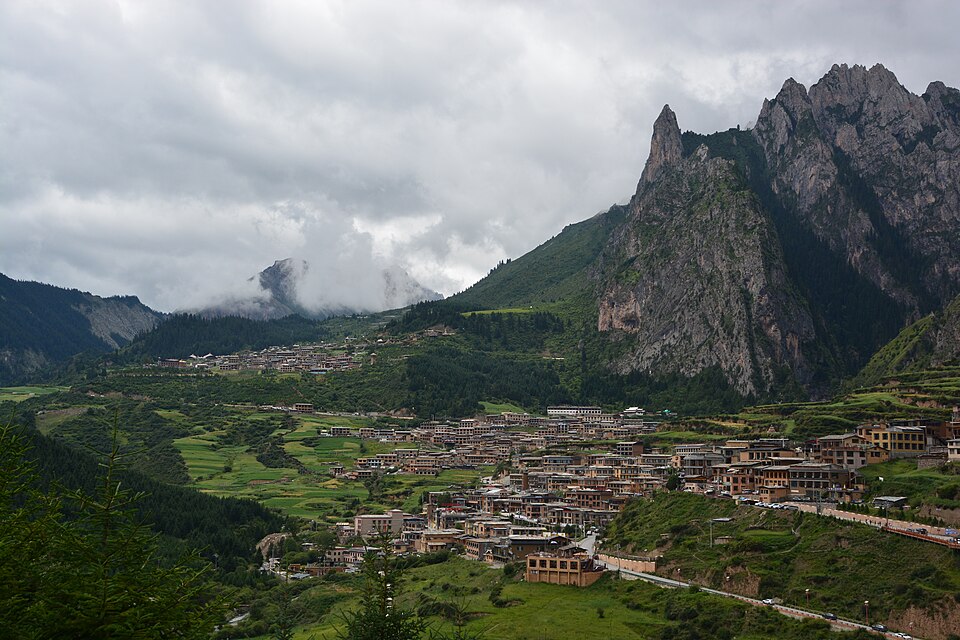 Un village aux toits sombres occupe le fond d'une vallée verte, dominé par une muraille de pitons rocheux dentelés que les nuages effleurent.