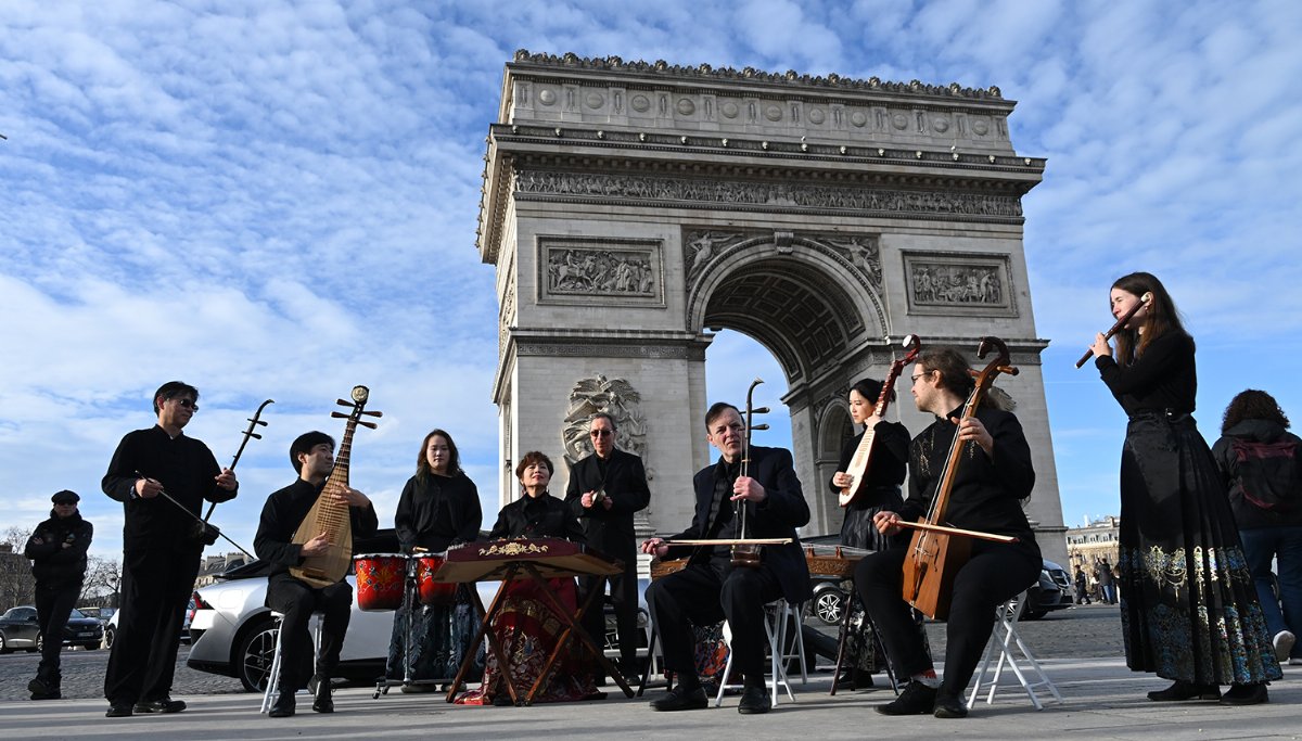 Une dizaine de musiciens en noir jouent en plein air devant l'Arc de Triomphe : deux vièles erhu, deux luths pipa, une cithare posée sur un tréteau, des percussions et une flûte traversière tenue debout à droite