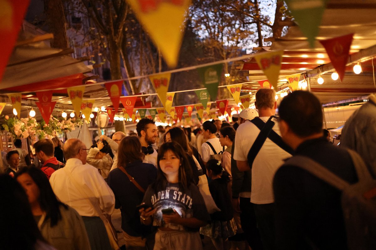 Photo de nuit du marché : une foule dense circule sous des guirlandes de fanions rouges et jaunes et des ampoules allumées, entre les toiles blanches des stands et les platanes du boulevard