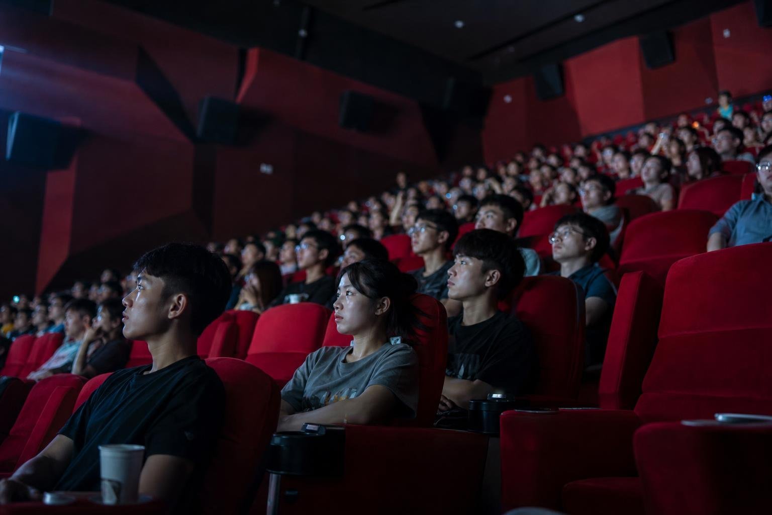Une salle de cinéma pleine, vue depuis le côté. Des rangées de fauteuils rouges occupées par de jeunes spectateurs qui regardent tous vers la gauche, le visage éclairé de bleu par l'écran hors champ. Au premier plan, un homme en tee-shirt noir avec un gobelet en carton posé dans l'accoudoir, une femme aux bras croisés. Les murs sombres portent des panneaux acoustiques rouges et des enceintes noires.