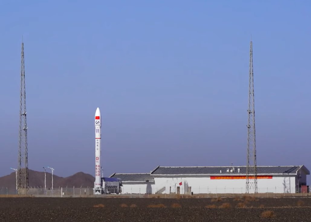Fusée Ceres-1 verticale sur son pas de tir, entre deux tours de service, dans un paysage désertique.