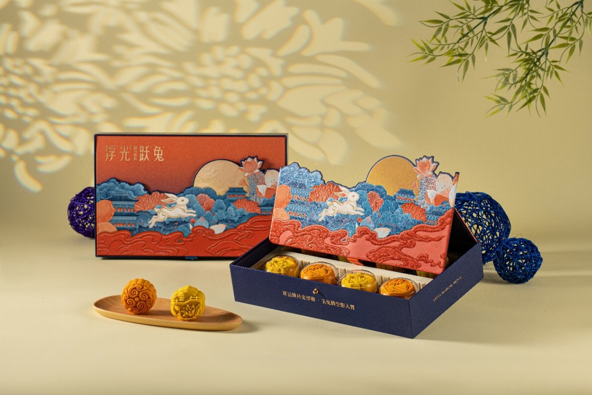 Coffret bleu nuit ouvert, couvercle orange illustré d'un lièvre blanc bondissant au-dessus de nuages et de pavillons, huit gâteaux dorés à l'intérieur