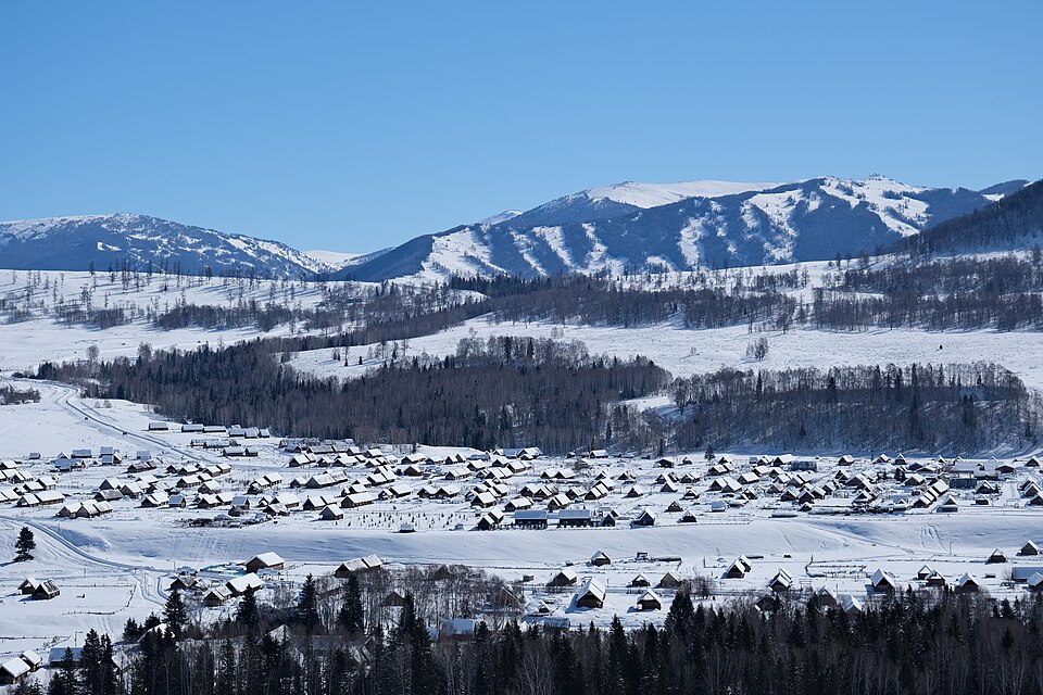 Un village de chalets de bois posé sur une plaine enneigée, cerné de sapins sombres et de croupes blanches ; au-dessus, un ciel d'hiver d'un bleu net, sans un nuage.