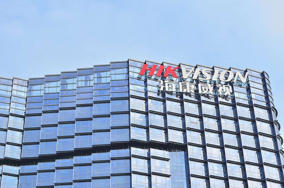 Enseigne HIKVISION en lettres rouges et blanches fixée au sommet d'un immeuble de bureaux entièrement vitré, sous un ciel bleu
