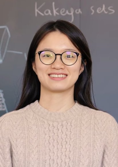 Portrait souriant de Hong Wang, lunettes rondes noires et pull beige torsadé, devant un tableau gris où sont écrits à la craie les mots Kakeya sets et deux schémas.