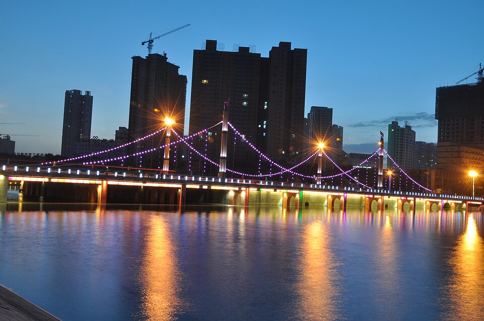 Un pont suspendu illuminé de guirlandes violettes franchit une rivière au crépuscule ; derrière lui se découpent les silhouettes noires des tours de la ville, et les lampes tracent de longues traînées orange sur l'eau.