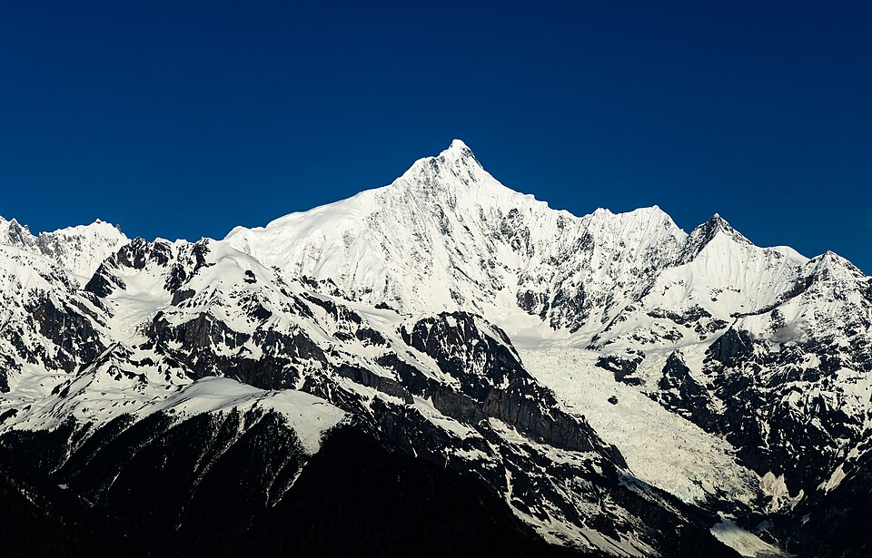 Une pyramide de neige et de glace se détache sur un ciel bleu presque noir ; à ses pieds, des arêtes rocheuses striées d'ombre descendent vers des pentes boisées très sombres.