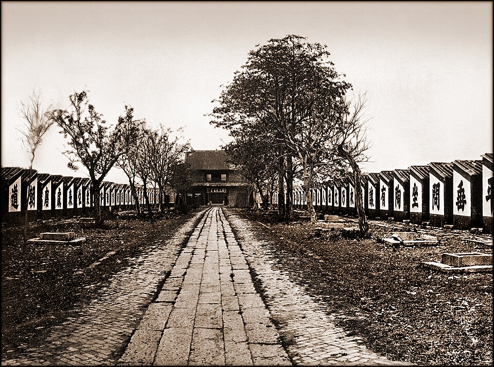 Photographie de 1873 : une allée pavée traverse la salle d'examen impérial de Canton, bordée de longues rangées basses de cellules dont chacune porte un grand caractère peint