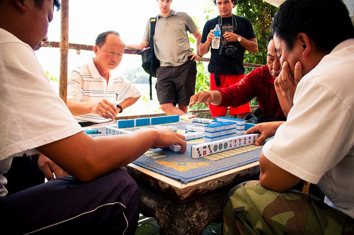 Quatre hommes jouent au mahjong autour d'une table de pierre sous un abri de bambou, les tuiles à dos bleu dressées en muraille devant eux ; deux voyageurs occidentaux regardent la partie en arrière-plan