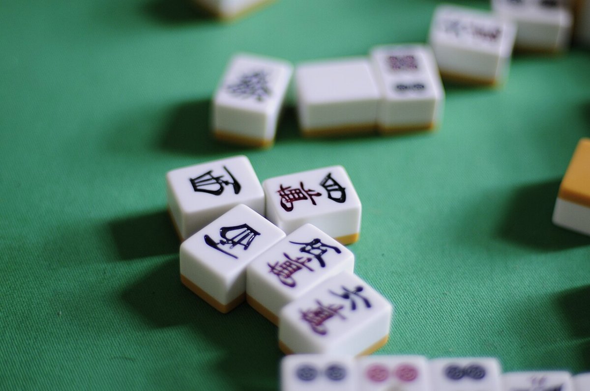 Gros plan sur des tuiles de mahjong posées sur un tapis vert : cinq tuiles gravées de caractères noirs et rouges au premier plan, une tuile à motif végétal derrière, et des tuiles à cercles bleus et roses au bord de l'image