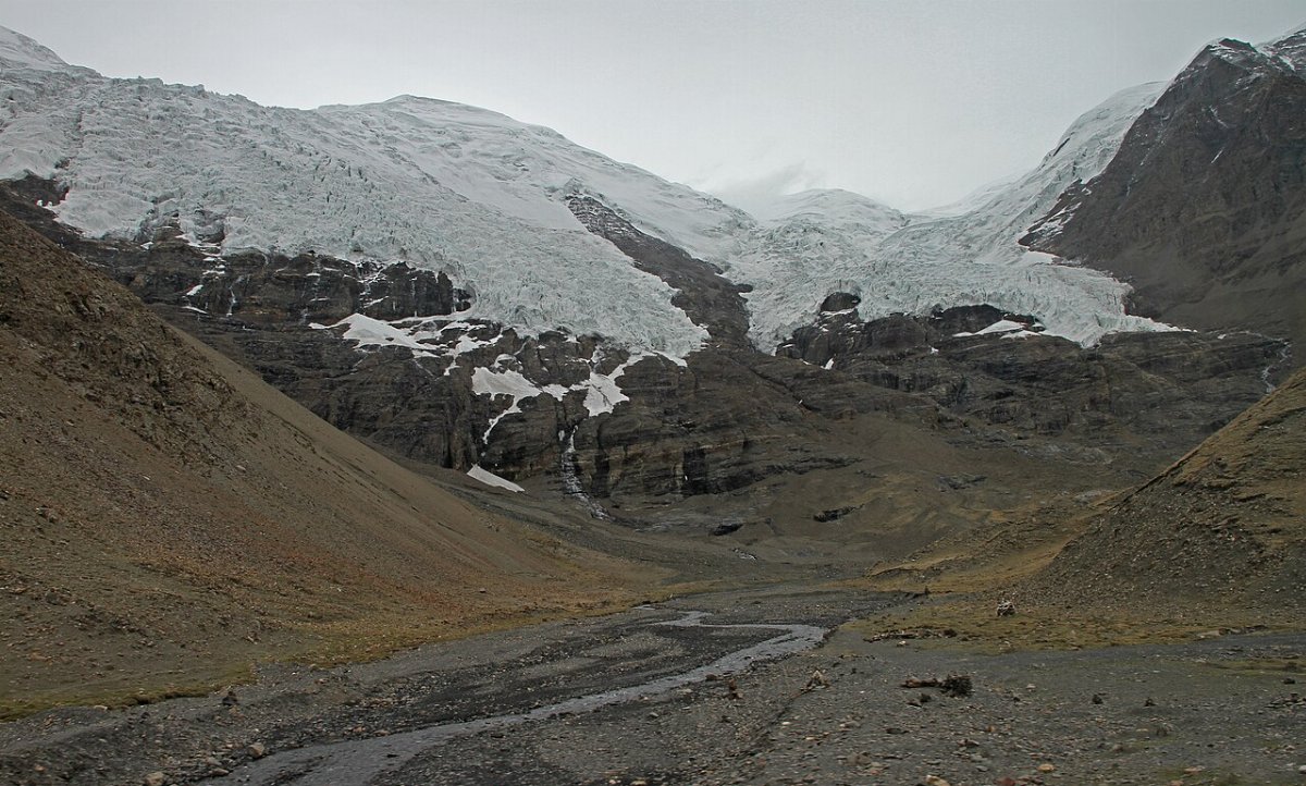 Langue glaciaire blanche et bleutée descendant une paroi rocheuse sombre jusqu'au lit caillouteux d'un torrent, sur le plateau tibétain