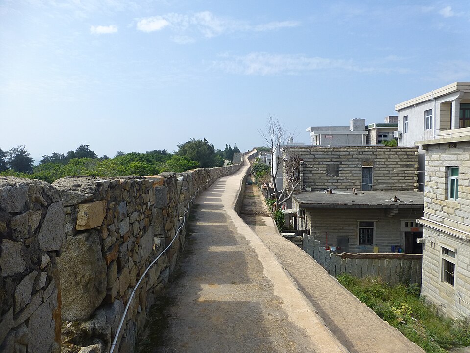Un chemin de ronde file au sommet d'un rempart de pierres grises assemblées à sec, qui ondule vers l'horizon ; à droite, en contrebas, les toits plats de maisons blanches, et au-dessus un ciel bleu pâle rayé de nuages fins.
