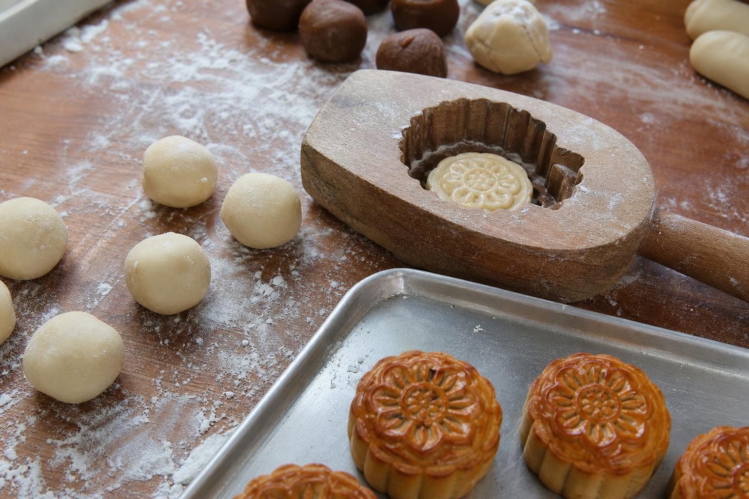 Sur un plan de travail fariné, des boules de pâte crue, un moule de bois sculpté contenant un gâteau de lune non cuit à motif de rosace, et trois gâteaux dorés sur une plaque métallique