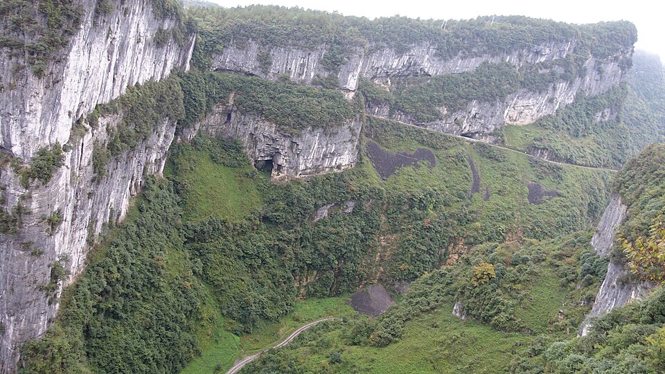 Des parois de calcaire clair, striées et couronnées de forêt, referment un gouffre immense où la végétation dévale les pentes ; un sentier étroit serpente tout au fond.
