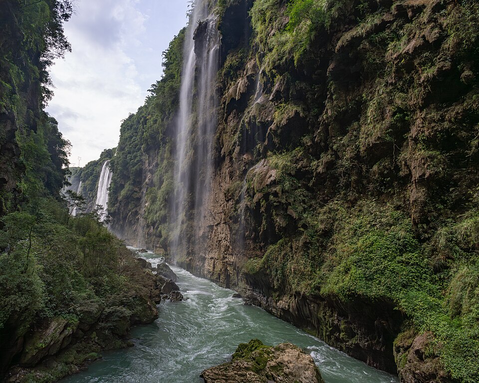 Une cascade tombe d'une paroi tapissée de mousses et de lianes dans une rivière vert jade qui bouillonne entre les rochers ; une seconde chute s'aperçoit plus loin dans le canyon.