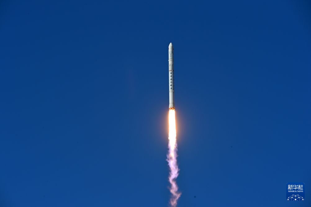 Fusée blanche au long corps effilé s'élevant dans un ciel bleu, laissant derrière elle une flamme orange et une traînée de fumée blanche.