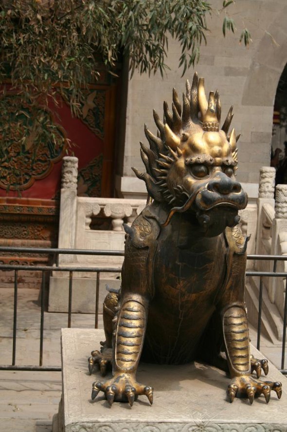 Statue de bronze représentant un animal fabuleux à tête de dragon, assis sur un socle de marbre devant une porte rouge sculptée