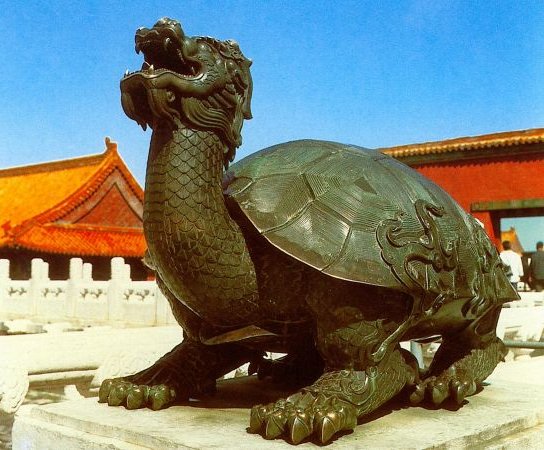 Tortue de bronze à tête de dragon cornue, gueule ouverte, posée sur un socle de marbre devant un bâtiment à toiture orangée