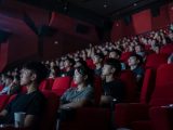 Une salle de cinéma pleine, vue depuis le côté. Des rangées de fauteuils rouges occupées par de jeunes spectateurs qui regardent tous vers la gauche, le visage éclairé de bleu par l'écran hors champ. Au premier plan, un homme en tee-shirt noir avec un gobelet en carton posé dans l'accoudoir, une femme aux bras croisés. Les murs sombres portent des panneaux acoustiques rouges et des enceintes noires.